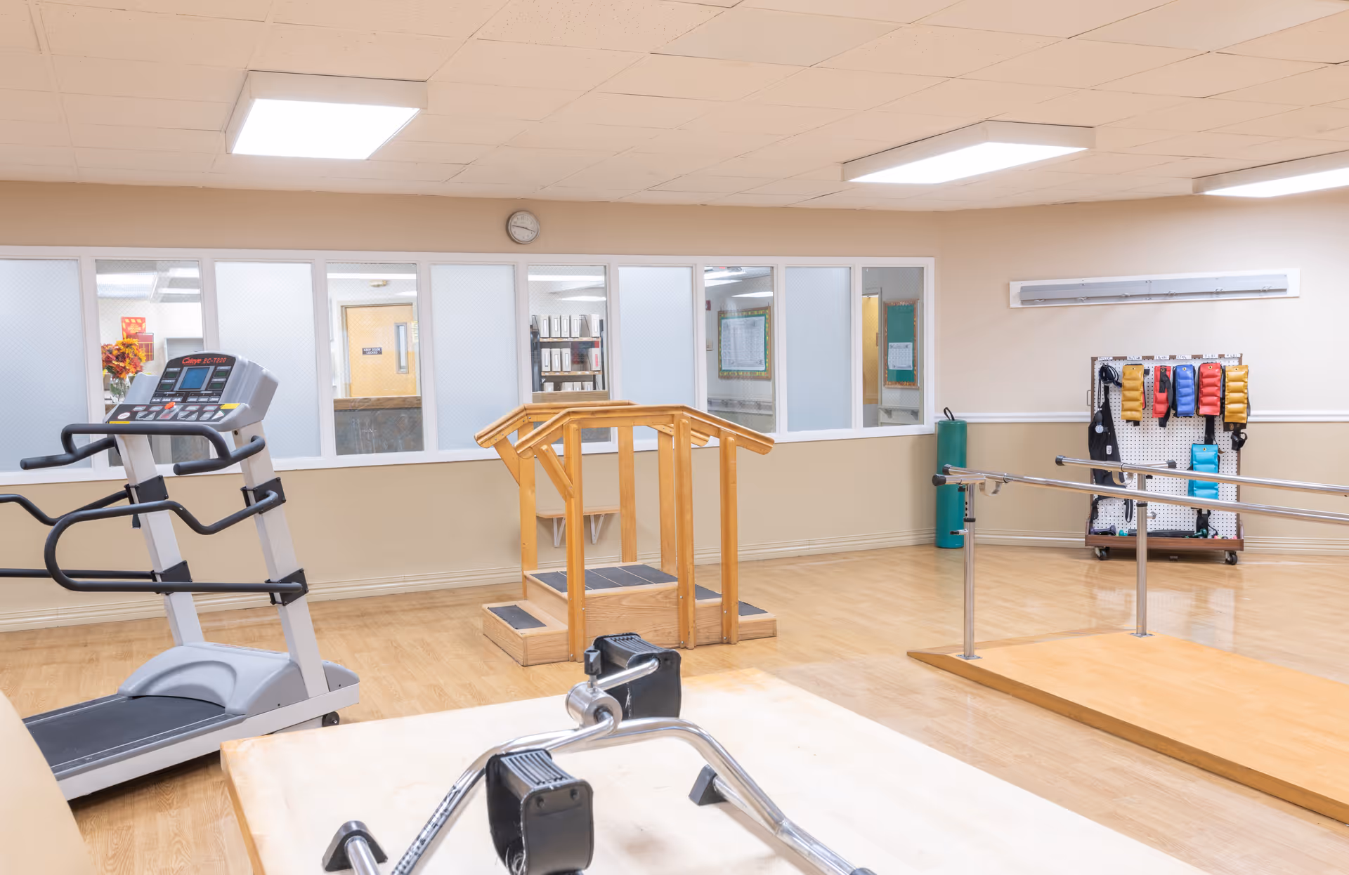 A rehabilitation therapy room with exercise equipment including a treadmill, parallel bars, a wooden step platform, and various weighted exercise bands hanging on a pegboard. The room has light-colored walls, a clock above a row of windows, and a wooden floor.
