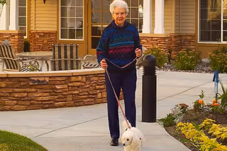 An elderly woman walking a small white dog on a leash along a paved pathway outside a senior living facility with landscaped gardens and outdoor seating areas visible.