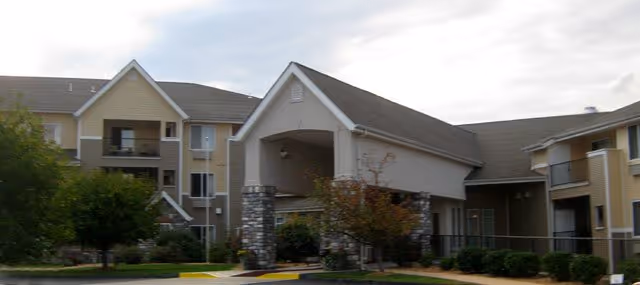 Exterior view of a senior living facility named College View Manor featuring a covered entrance supported by stone pillars, surrounded by landscaped bushes and trees, with multiple residential buildings in the background under a cloudy sky.