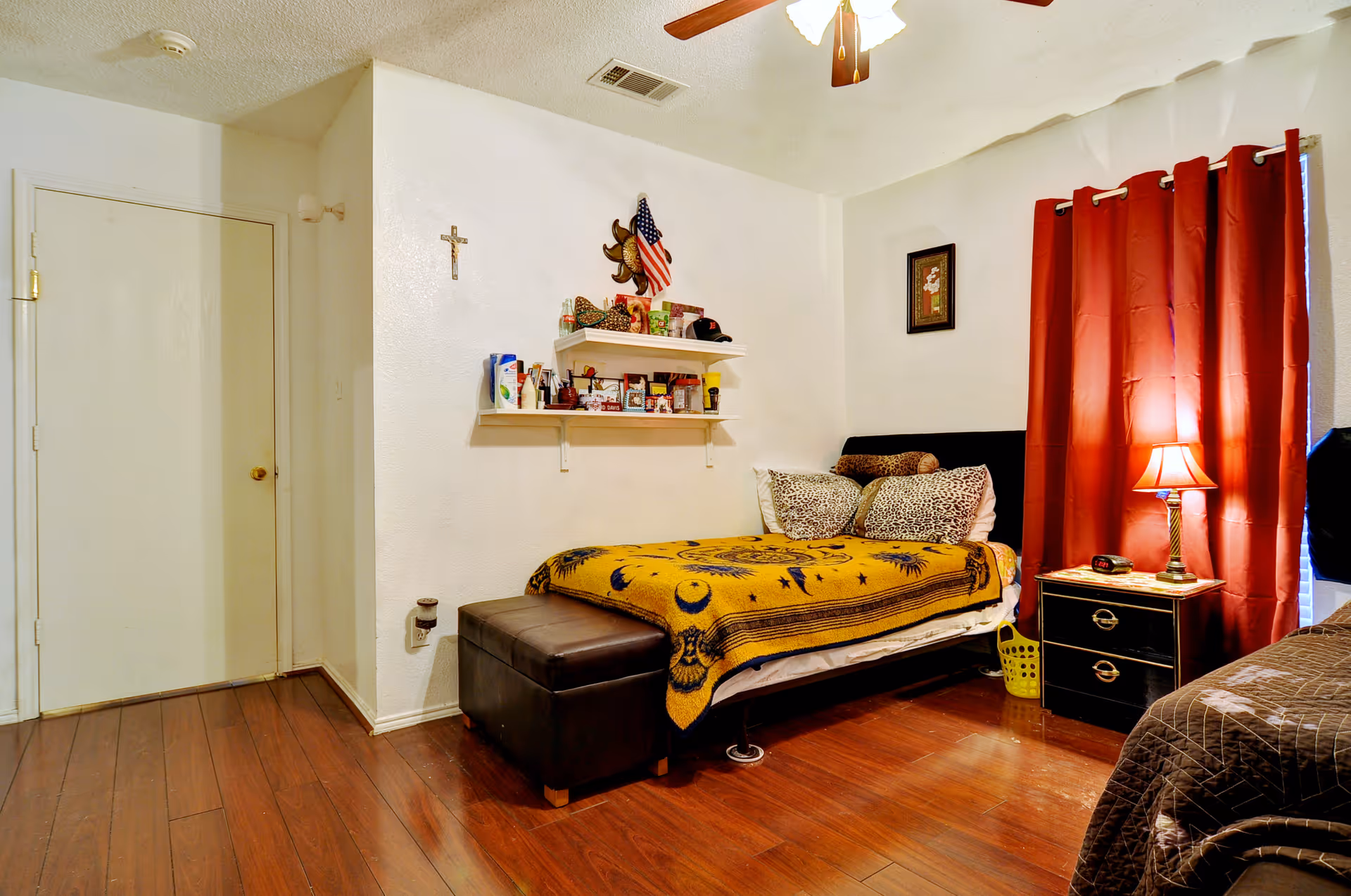 Bedroom with a single bed covered by a yellow patterned blanket, red curtains, a nightstand with a lamp, and wooden flooring.