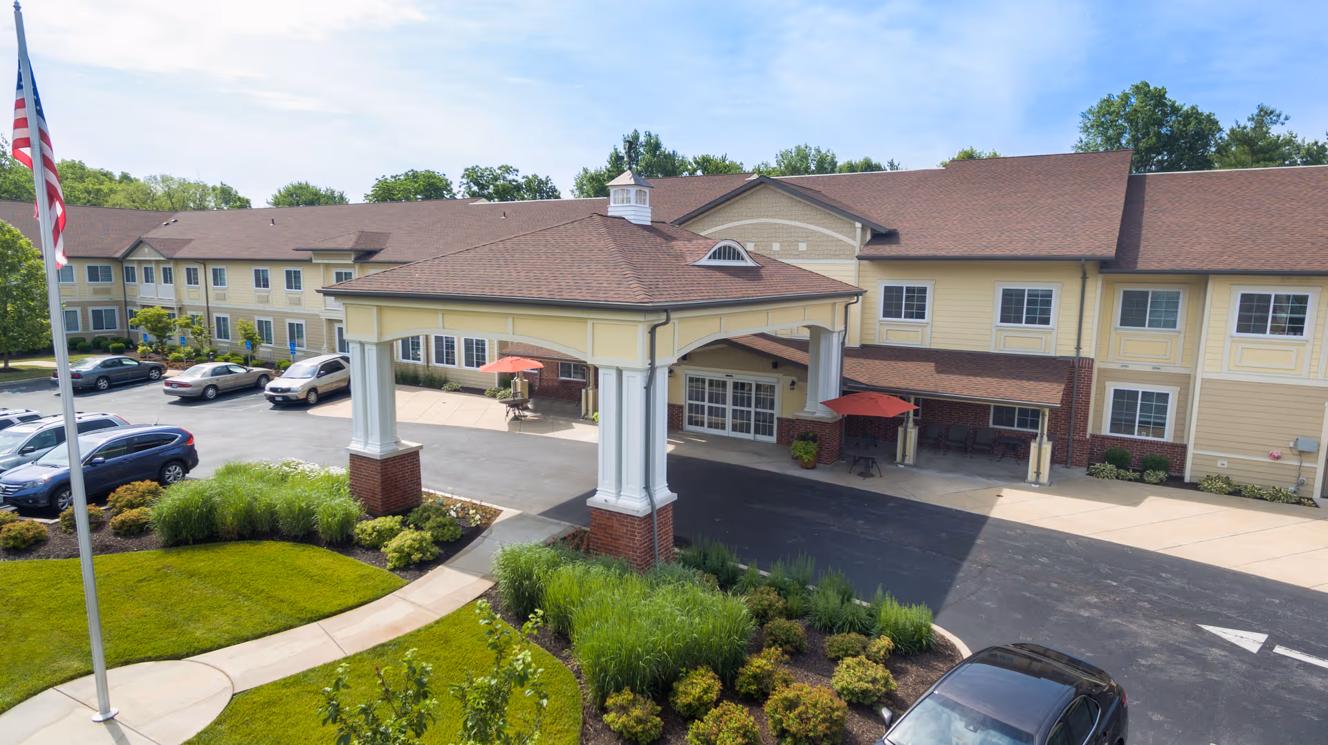 Exterior view of Southview Assisted Living & Memory Care facility showing a two-story building with beige siding and a brown roof. There is a covered entrance with white columns and brick bases, a driveway with parked cars, landscaped greenery, and an American flag on a flagpole.
