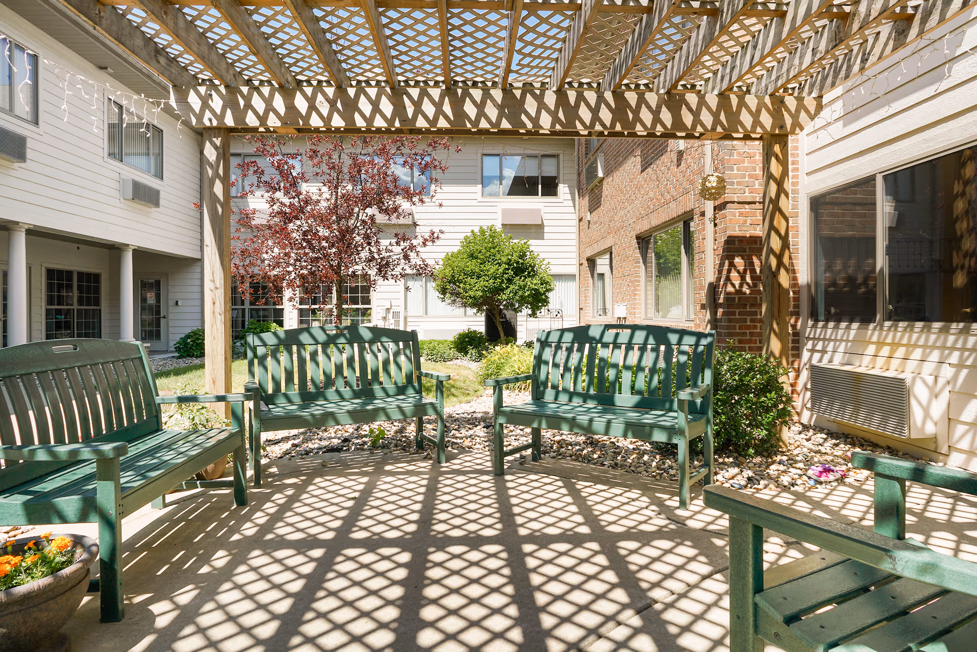 Outdoor seating area with green benches under a wooden lattice pergola casting patterned shadows on the ground. Surrounding the area are buildings with white and brick exteriors, windows, and some greenery including a small tree and bushes.