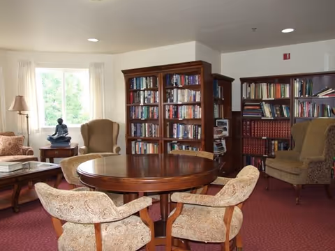Well-lit community reading room with a round wooden table surrounded by upholstered chairs, bookshelves, and armchairs by a window.
