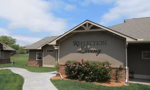 Front exterior of Reflection Living facility with sign, shrubs, and a curved walkway.