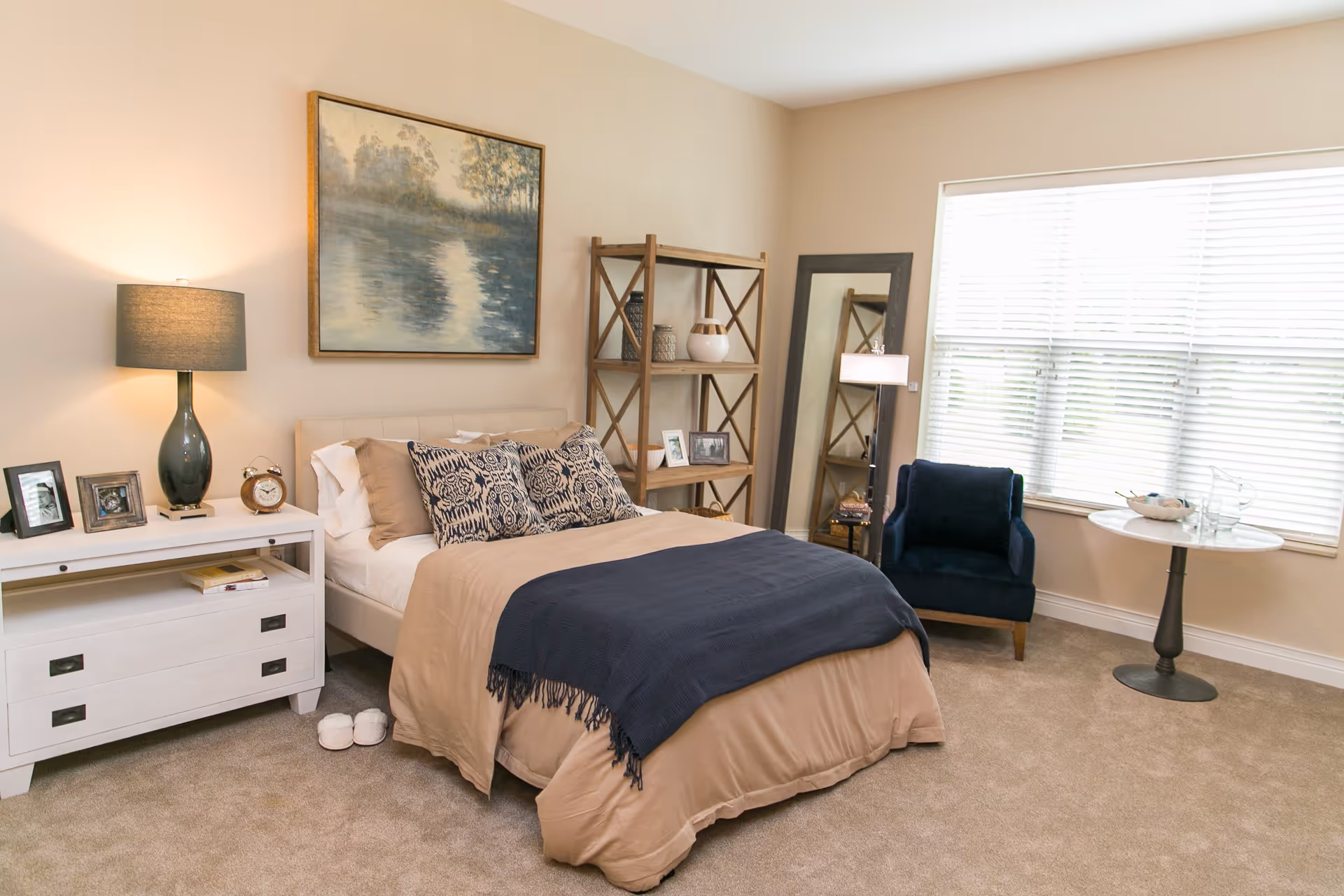 A cozy bedroom with a beige bedspread and dark blue throw blanket on a bed with patterned pillows. Next to the bed is a white nightstand with a lamp, clock, and framed photos. A wooden shelving unit with decorative items and a large mirror stand against the wall. A dark blue armchair and a small round table with glassware are placed near a large window with blinds. The room has beige carpet and walls, and a framed painting of a serene water scene hangs above the bed.