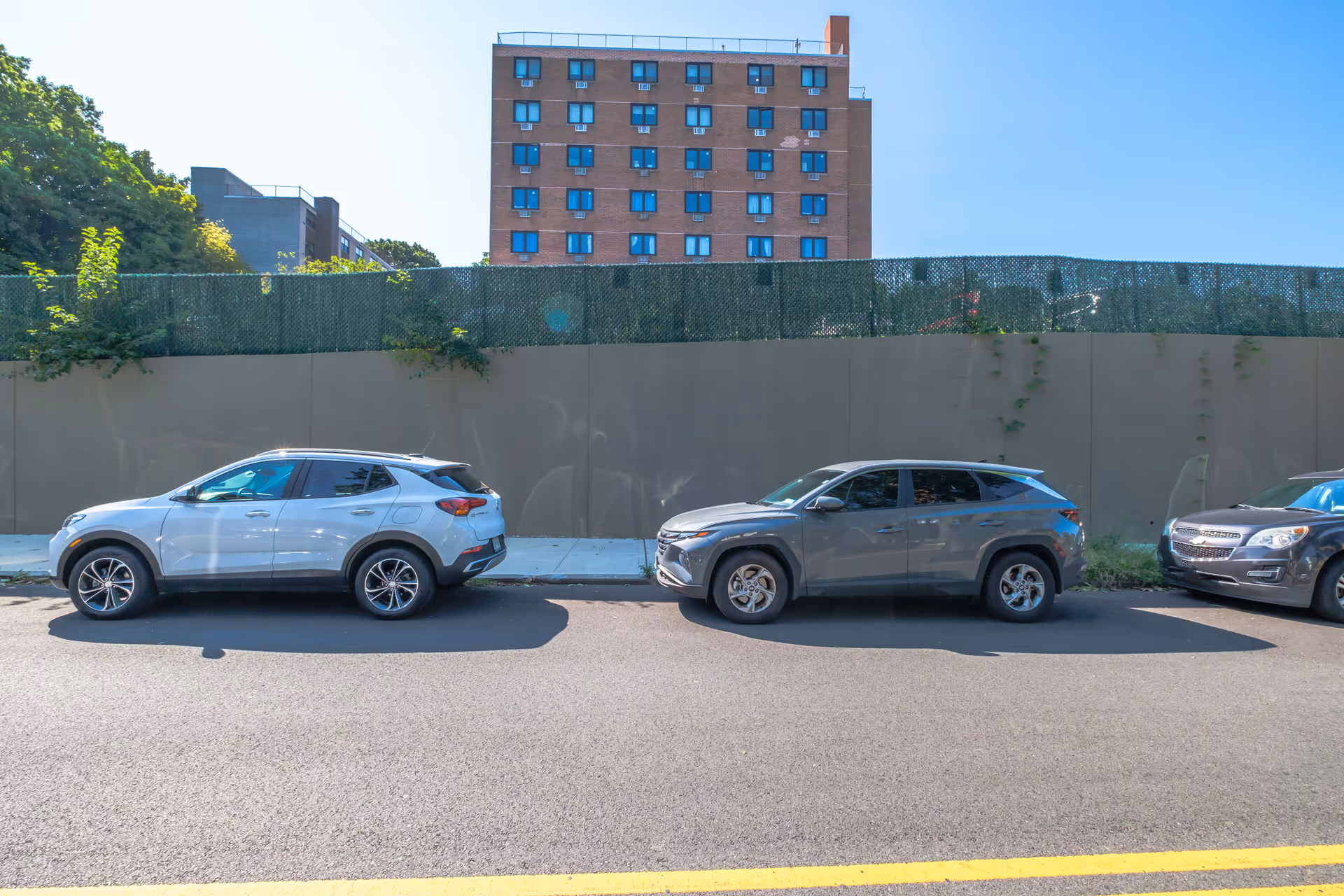 Three cars parked along a street in front of a tall brick building with multiple windows. A green mesh fence and a beige wall separate the street from the building. Trees and clear blue sky are visible in the background.