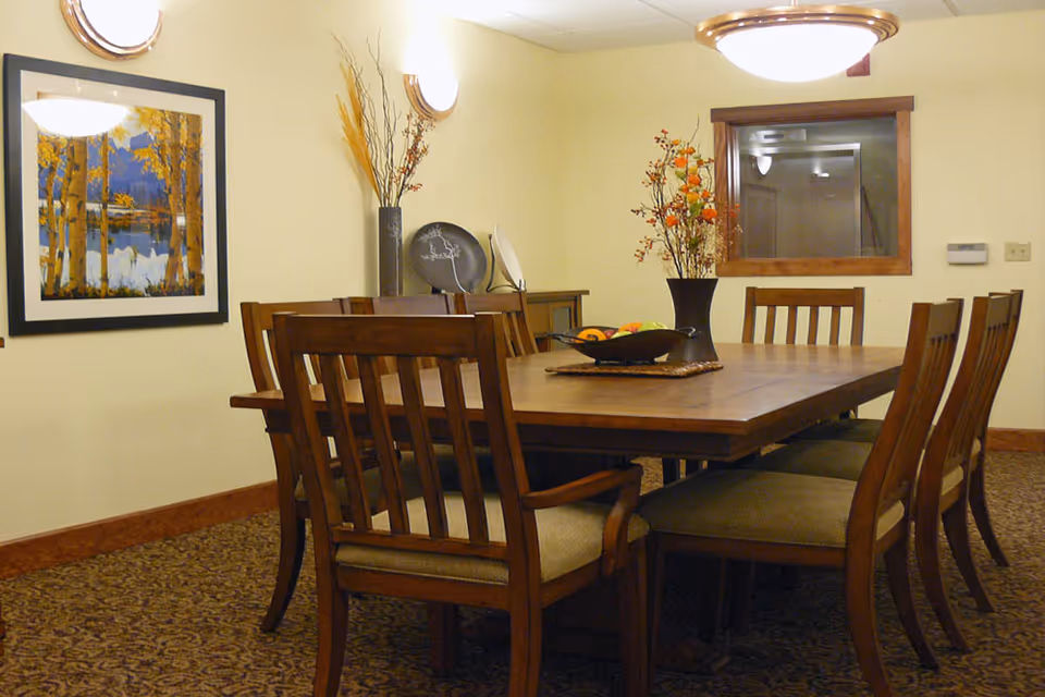 A dining room with a large wooden table surrounded by eight wooden chairs with cushioned seats. The table has a decorative centerpiece with a bowl of fruit and a vase with orange and red flowers. The room has beige walls, a framed painting of a forest scene, two wall-mounted lights, a window with wooden trim, and a ceiling light fixture.