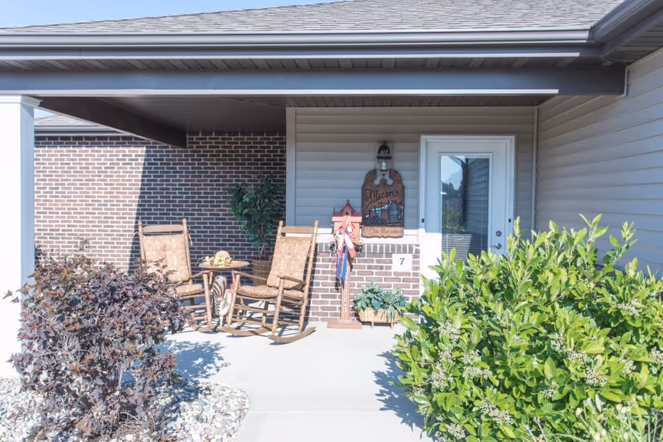 Front porch entry with two wooden rocking chairs and a small table beside a glass door under a covered overhang.