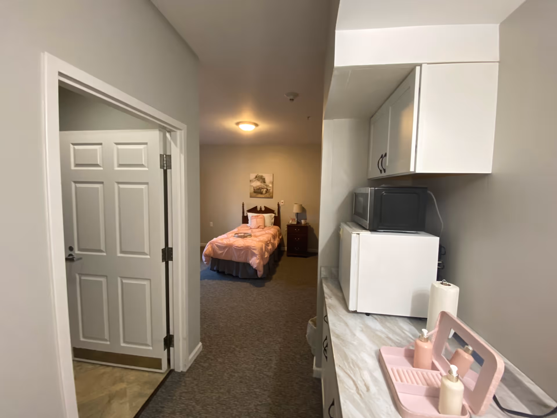 View down a hallway in a senior living facility room showing a small kitchenette area with a microwave, mini fridge, paper towel holder, and soap dispensers on a pink tray. At the end of the hallway is a bedroom with a single bed covered in a pink comforter, a nightstand with a lamp, and a framed picture on the wall.