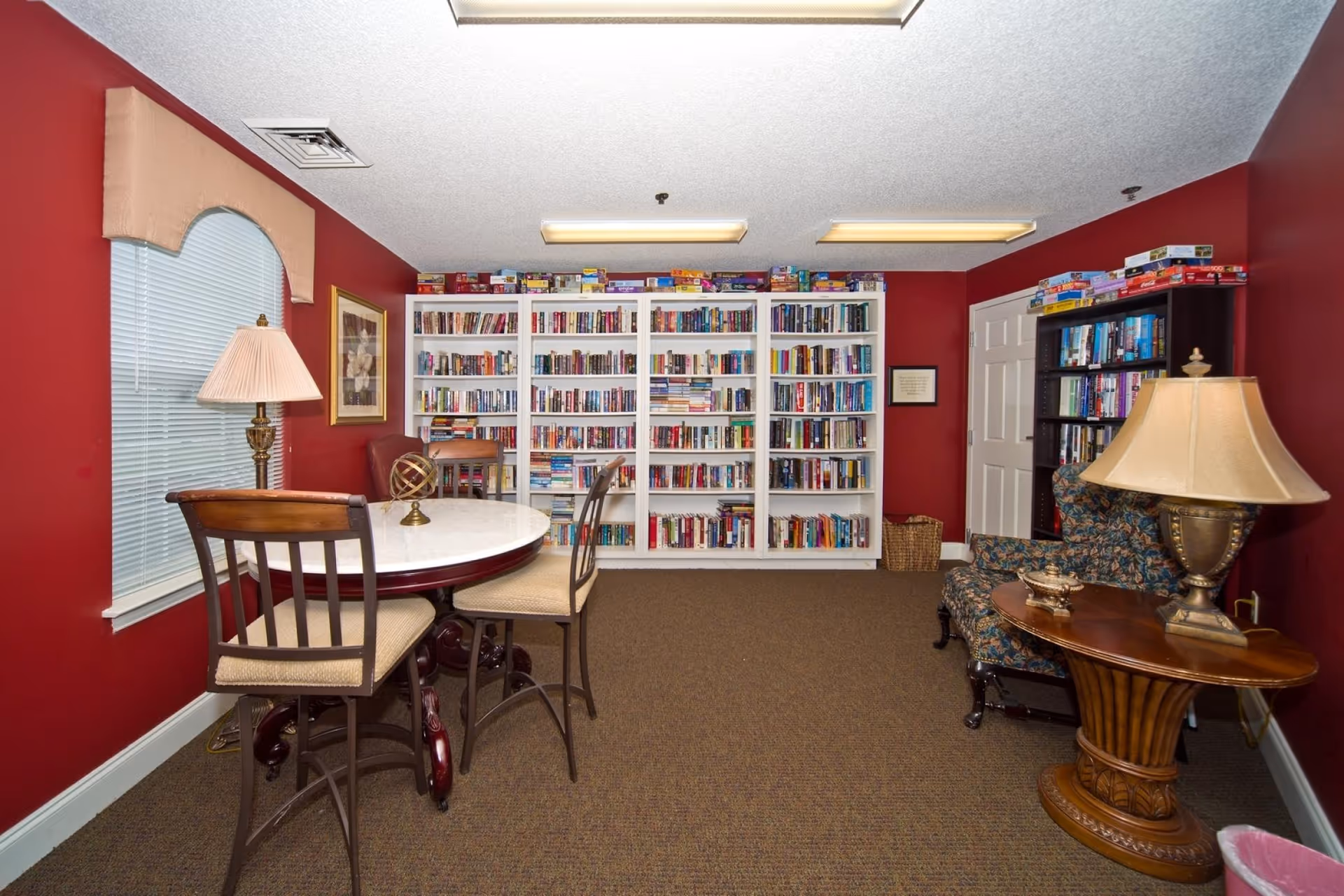 A cozy reading room with red walls featuring a round table with three chairs, a large white bookshelf filled with books and board games on top, a window with blinds and a valance, a framed picture on the wall, a floral upholstered armchair, a wooden side table with a lamp, and a carpeted floor.