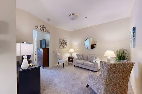 A cozy living room with a beige sofa, two patterned armchairs, a round mirror on the wall, a side table with a lamp, and a decorative plant. The room has light-colored walls and carpeted flooring. A doorway leads to another room with a dresser and a TV.