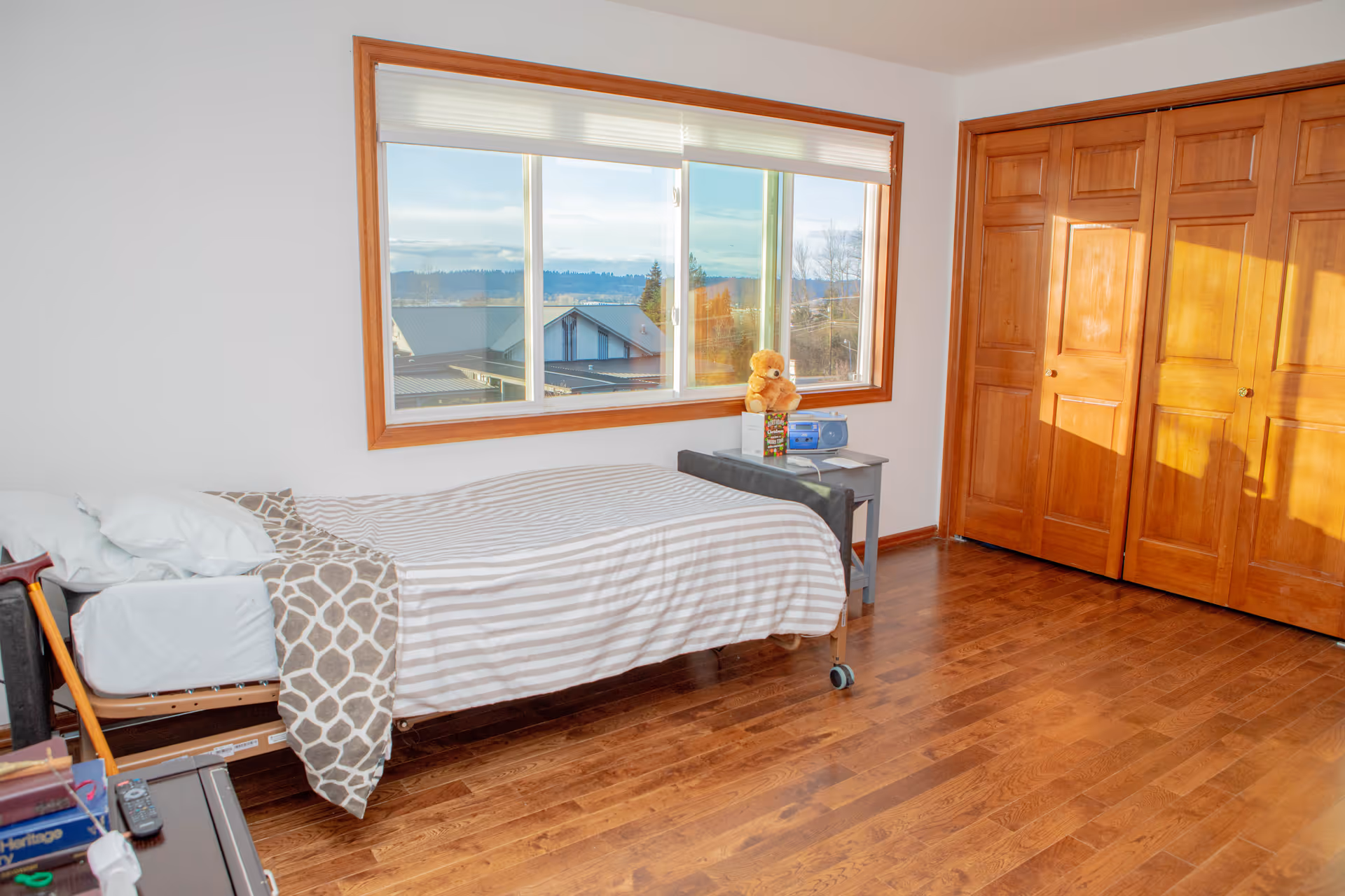 A bright bedroom with a single bed covered in striped bedding and a patterned blanket. There is a small gray nightstand next to the bed with a teddy bear, a radio, and some books on it. The room has wooden floors, a large window with a view of rooftops and trees outside, and wooden closet doors on the right side.