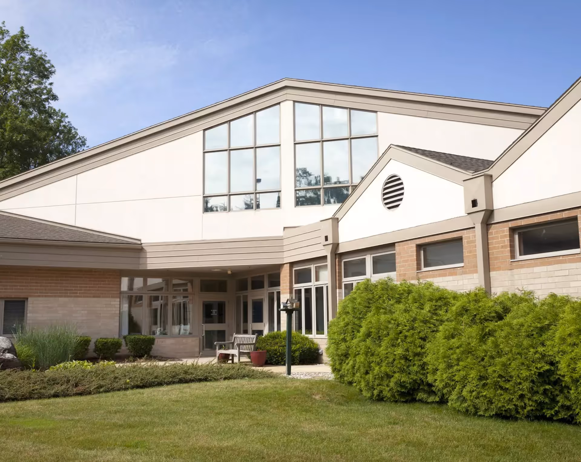 Exterior view of StoryPoint Kalamazoo (Bronson Place) building with large windows, beige and brick walls, green bushes, and a well-maintained lawn under a clear blue sky.