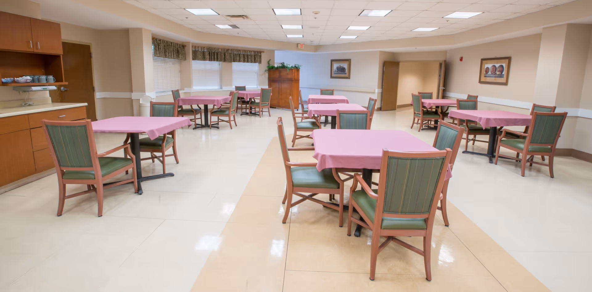 A spacious dining room with several square tables covered with pink tablecloths, each surrounded by green cushioned chairs with wooden frames. The room has a tiled floor, beige walls, ceiling lights, and windows with blinds and valances. There is a wooden cabinet and framed artwork on the walls.