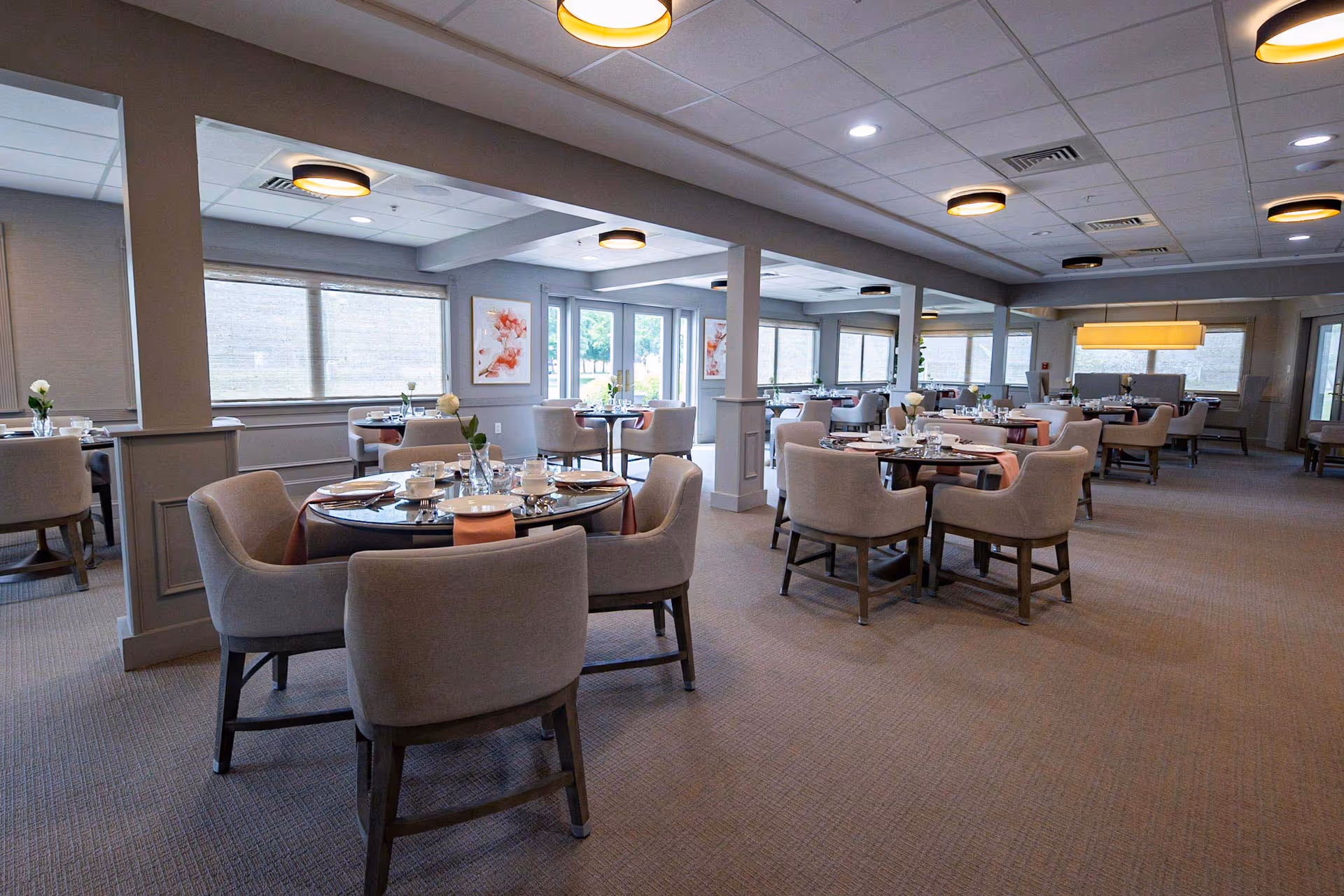 A spacious dining room with multiple round tables set with plates, cups, and silverware. Each table has a small vase with a white flower. The room has large windows with blinds, soft lighting from ceiling fixtures, and neutral-colored walls and carpet. Artwork is displayed on the walls, and there are comfortable upholstered chairs around each table.