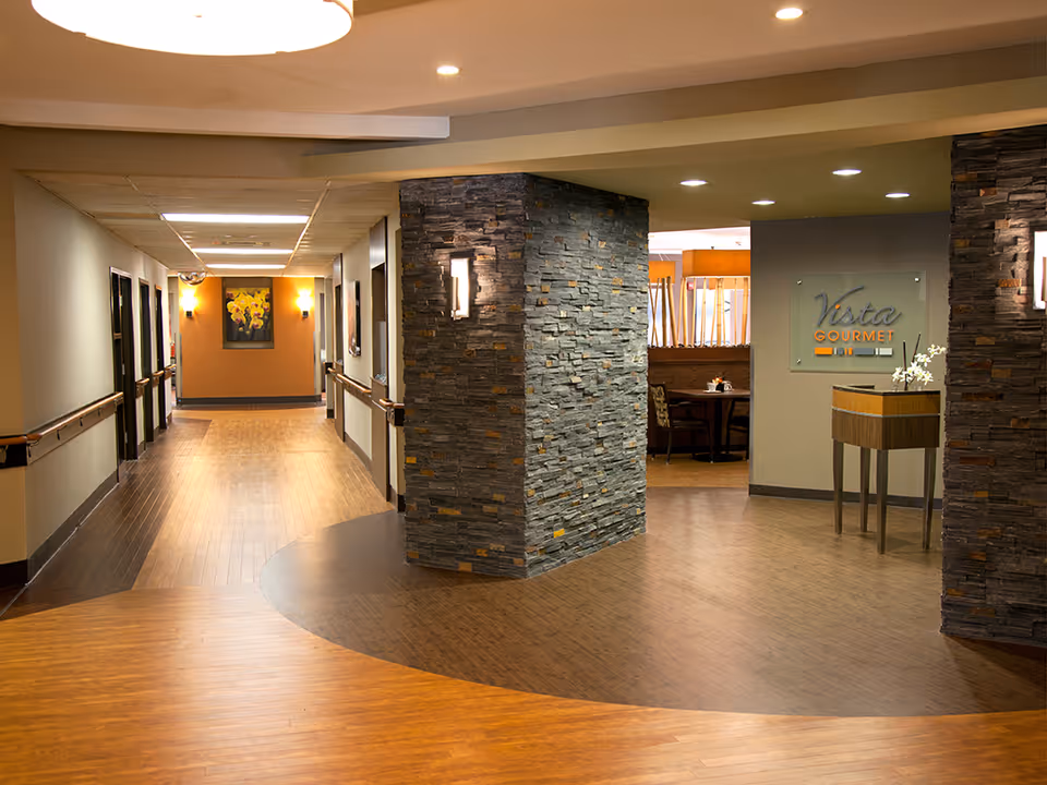 Interior view of a senior living facility hallway with wooden floors and stone pillars. The hallway leads to a dining area named 'Vista Gourmet' with tables and chairs. The walls are decorated with framed artwork and wall sconces provide lighting.
