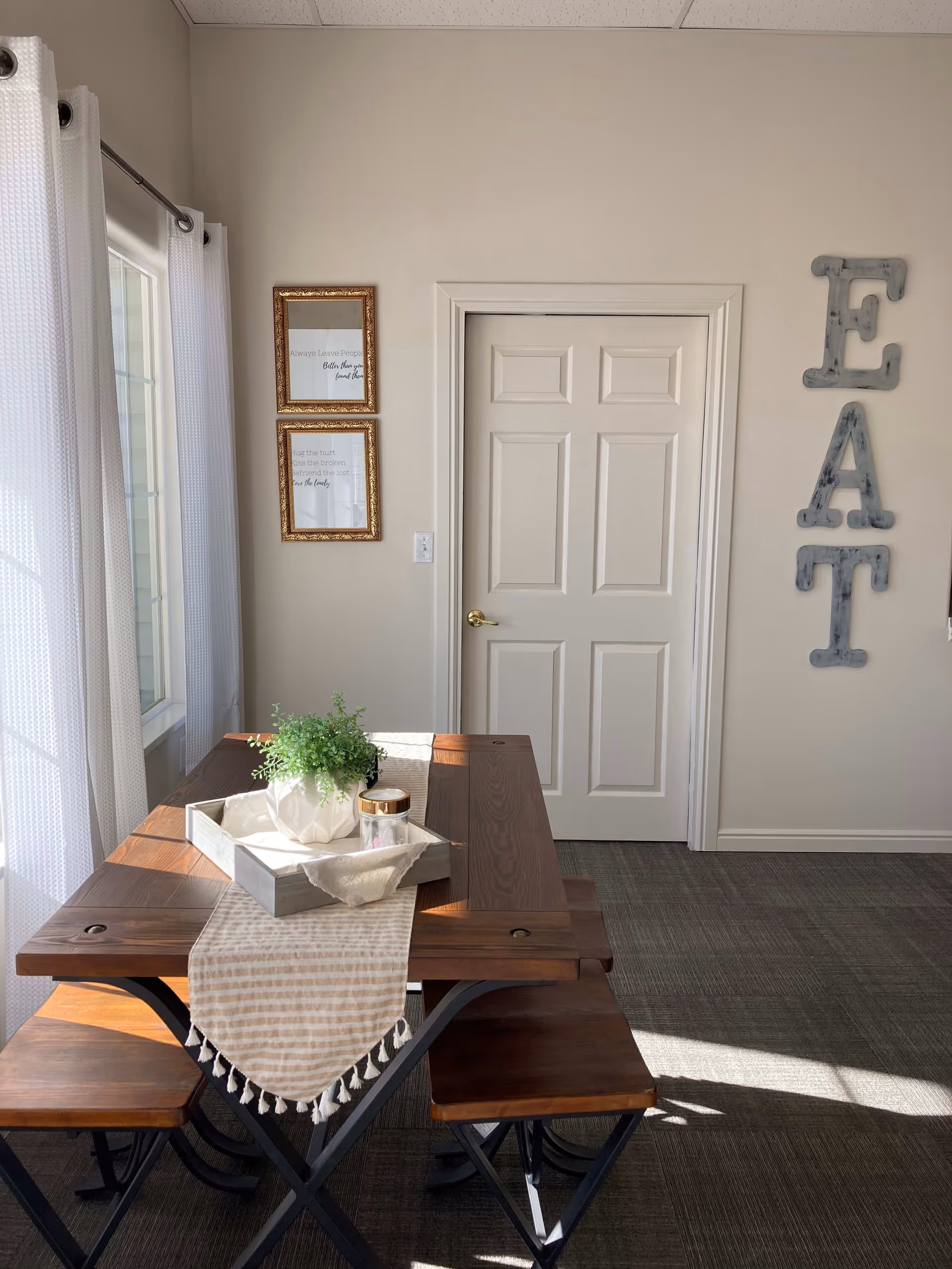 A small dining area with a wooden table and matching benches. The table has a beige striped runner with tassels and a tray holding a white vase with green plants and a small jar. To the left, there is a window with white curtains allowing sunlight to brighten the room. On the wall, there are two framed quotes and large metal letters spelling 'EAT'. A white door with a gold handle is centered on the back wall.