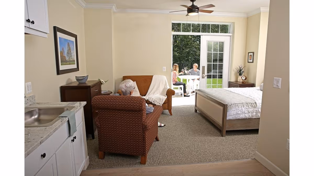 A senior living facility room with a bed, armchair, and small kitchenette area. An elderly person is seated in the armchair facing a glass door that opens to an outdoor patio where two people are sitting at a table.