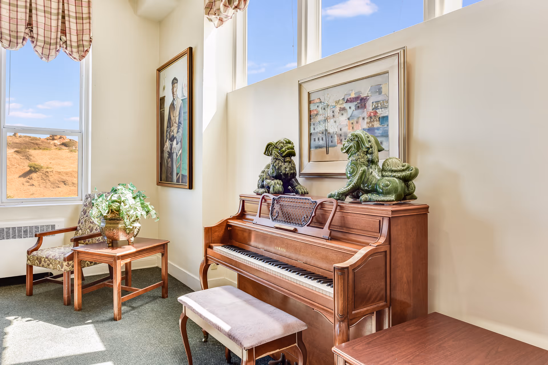 A cozy corner of a room featuring a wooden upright piano with two green decorative statues on top. Above the piano hangs a framed painting of a colorful village. To the left, there is a wooden chair with floral upholstery next to a small wooden table holding a potted plant. The room has large windows with plaid valances, allowing natural light to brighten the space and offering a view of a dry, hilly landscape outside.