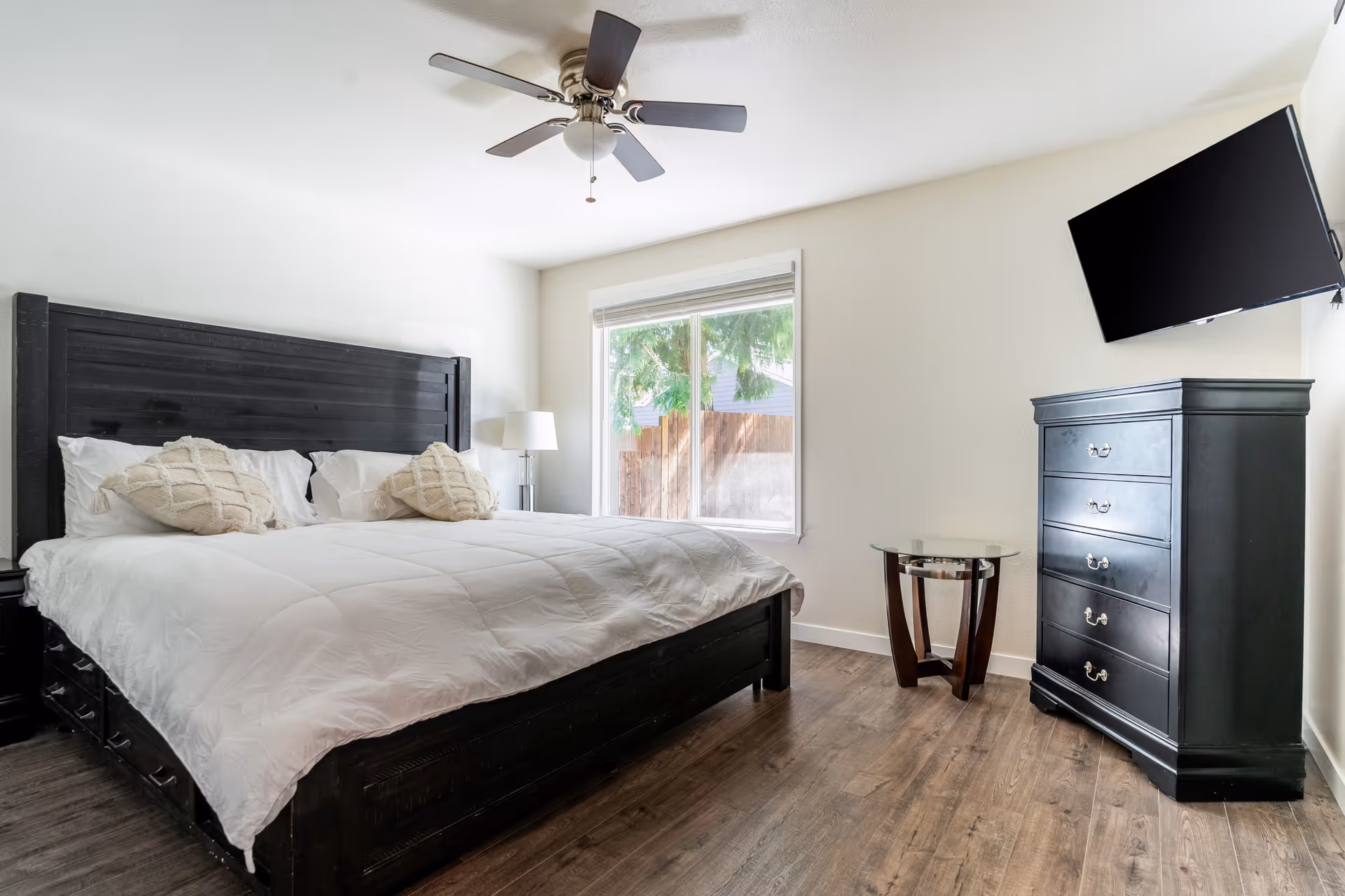 Bright bedroom with a large dark wood bed dressed in white bedding, ceiling fan, window, dresser, wall-mounted TV and a small side table.