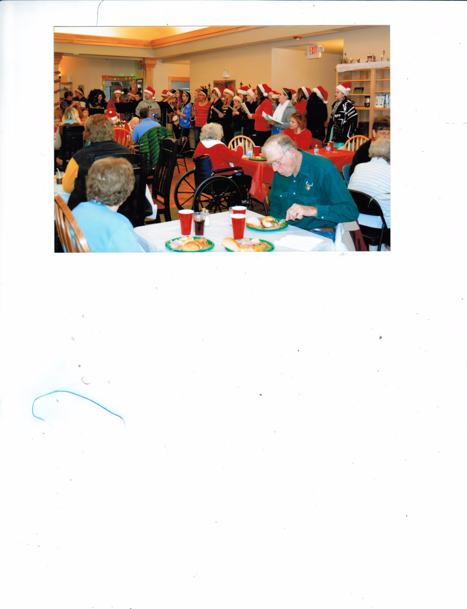 A group of elderly people seated at tables with food and drinks in a dining area. In the background, a choir wearing Santa hats is performing, creating a festive atmosphere. Some residents are in wheelchairs, and the room is decorated for the holiday season.