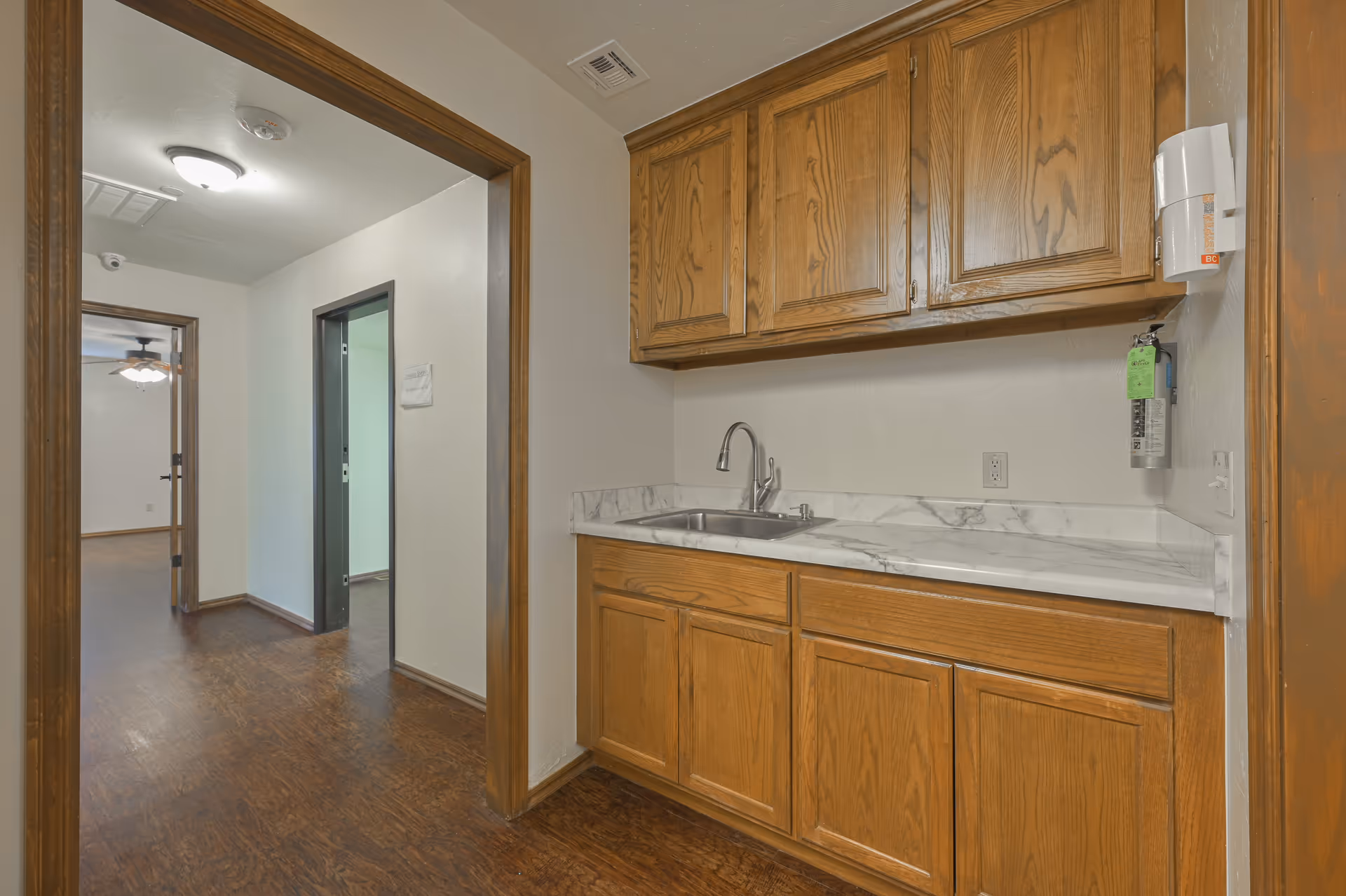 A small kitchenette with oak cabinets, a marble-look countertop and sink beside a hallway leading to adjacent rooms.