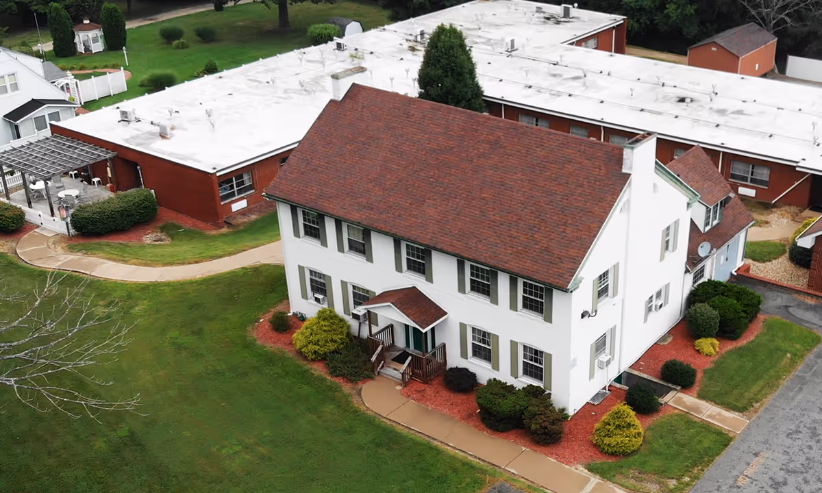 Aerial view of Havencrest Healthcare and Rehabilitation Center showing a large white two-story building with a red roof surrounded by green lawns, bushes, and paved walkways. Adjacent to it is a long single-story brick building with a white roof and a small gazebo in the background.