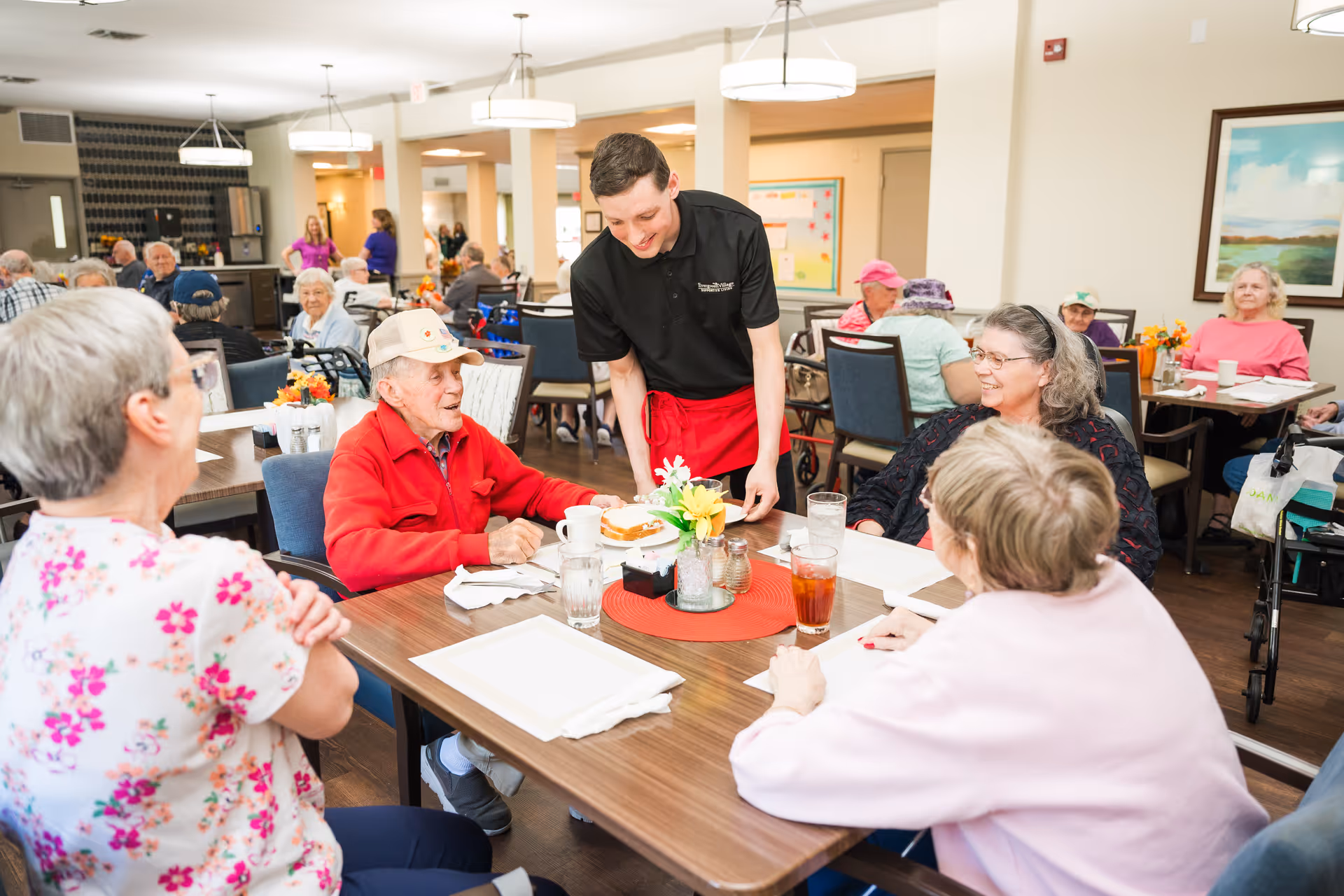 A server brings a meal to a table of smiling elderly residents dining together in a senior living facility dining room.