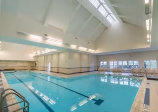 Indoor swimming pool with clear blue water, surrounded by tiled flooring and white walls. The ceiling is high with skylights allowing natural light to enter. There are several windows along one side and pool ladders at the edges.