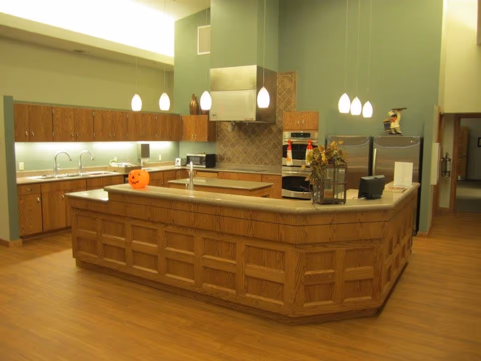 A spacious kitchen area with wooden cabinets and a large central island with a countertop. The kitchen features two sinks, stainless steel appliances including ovens and refrigerators, pendant lights hanging from the ceiling, and a small pumpkin decoration on the island. The walls are painted green and the floor has a wood finish.
