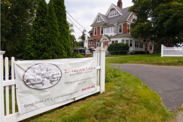 Front exterior of a large Victorian-style house with a white fence and a banner reading 'Riverview Lodge Residential Care Home' on the lawn.
