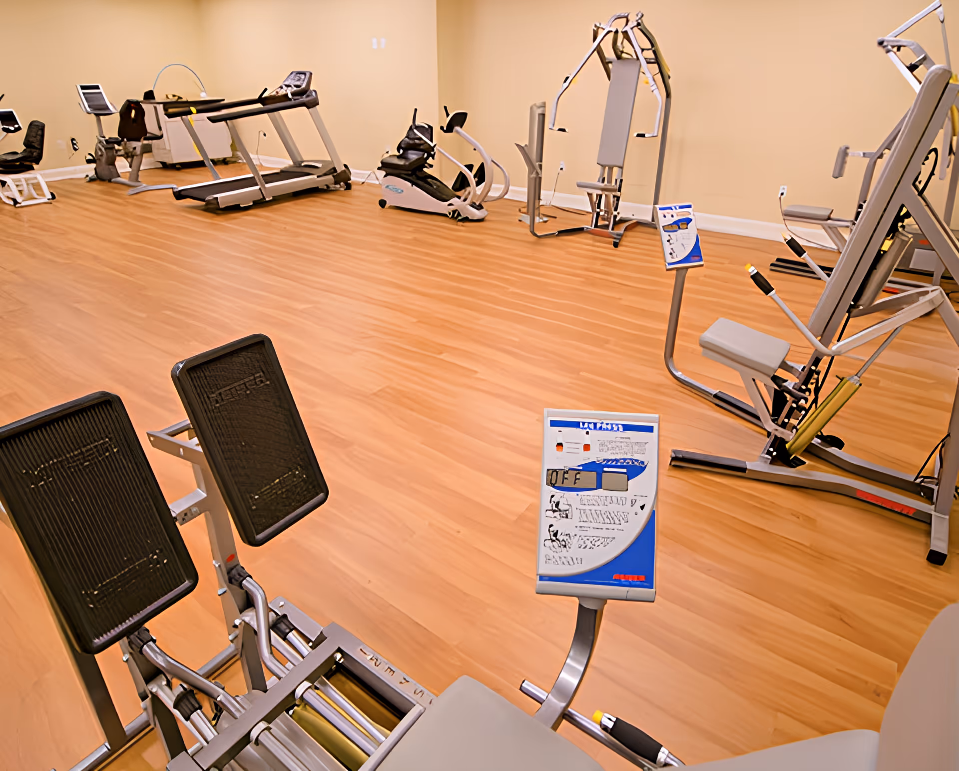A fitness room with various exercise equipment including a treadmill, stationary bike, and strength training machines on a wooden floor with beige walls.