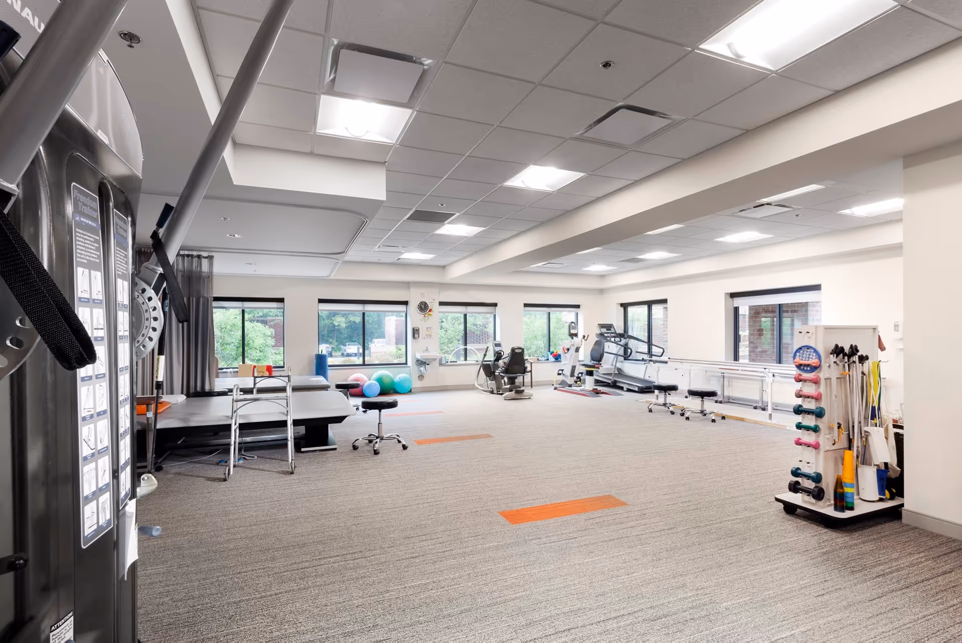 A spacious physical therapy or rehabilitation room with exercise equipment including a treadmill, stationary bike, therapy tables, exercise balls, weights, and walking aids. The room has large windows letting in natural light and a carpeted floor with orange accents.