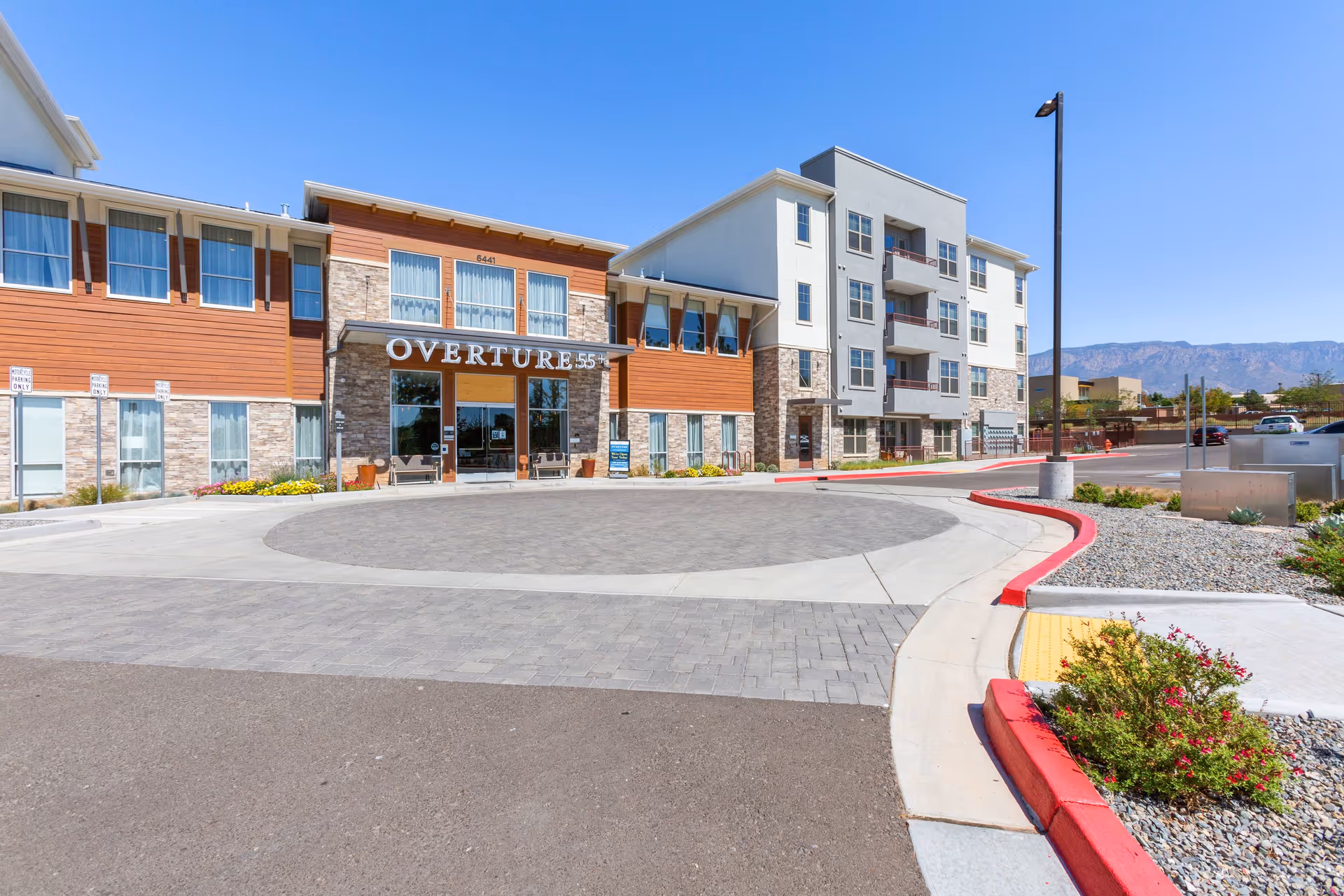 Front entrance of the Overture 55+ senior living building with a circular driveway, landscaping, and a multi-story facade under a clear blue sky.