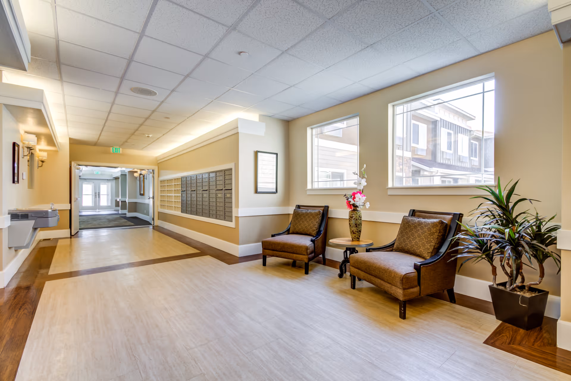 A bright hallway in a senior living facility with two cushioned chairs and a small table with a vase of flowers near two large windows. There are mailboxes mounted on the wall and a water fountain on the left side. The floor has a wood and tile pattern, and the hallway leads to a door with glass panels.