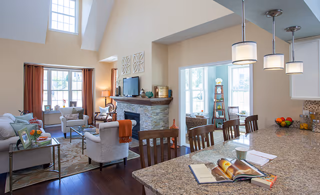 A bright and spacious living room and kitchen area in a senior living facility. The living room features a stone fireplace with a TV mounted above it, comfortable seating including a sofa and armchairs, and large windows with brown curtains letting in natural light. The kitchen area has a granite countertop with three pendant lights hanging above, wooden chairs, and an open book and notebook on the counter. The room has high ceilings and a warm, inviting atmosphere.