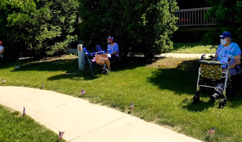 Two elderly individuals sitting outside on a grassy area near a sidewalk, each with a walker. They are wearing face masks and casual clothing. Small American flags are placed along the edge of the sidewalk. There are bushes and a building in the background.