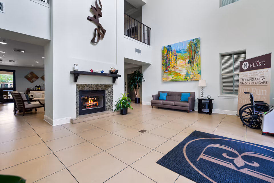 A spacious and bright senior living facility lobby with a tiled floor, a modern fireplace with a decorative mantle, a gray sofa with blue cushions, a colorful abstract painting on the wall, a potted plant, a side table with a lamp, and a wheelchair near a banner for The Blake. The area has high ceilings and an open balcony above.