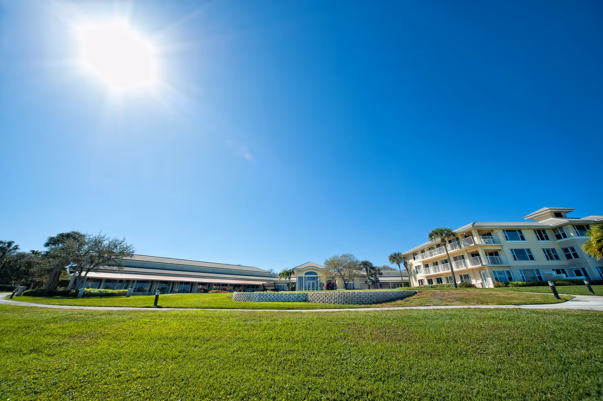 Wide exterior view of Sandhill Cove Retirement Living facility under a bright blue sky with the sun shining. The image shows a large green lawn in the foreground, a circular stone planter with plants, and two main buildings with multiple windows and balconies surrounded by palm trees and other greenery.