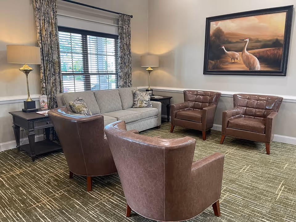 A cozy sitting area with a beige sofa, two brown leather armchairs, and two matching side tables with lamps. The room has a large window with patterned curtains and a painting of two cranes on the wall above the armchairs. The carpet is green with a linear pattern.