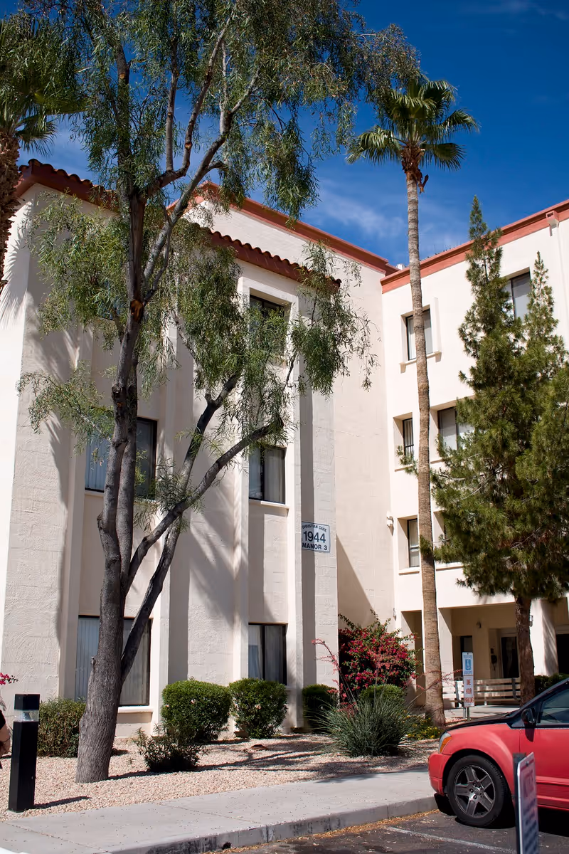 Exterior view of a three-story beige building with red roof trim, surrounded by trees and shrubs under a clear blue sky. A red car is parked in front of the building, which has a sign reading '1944 Manor 3'.