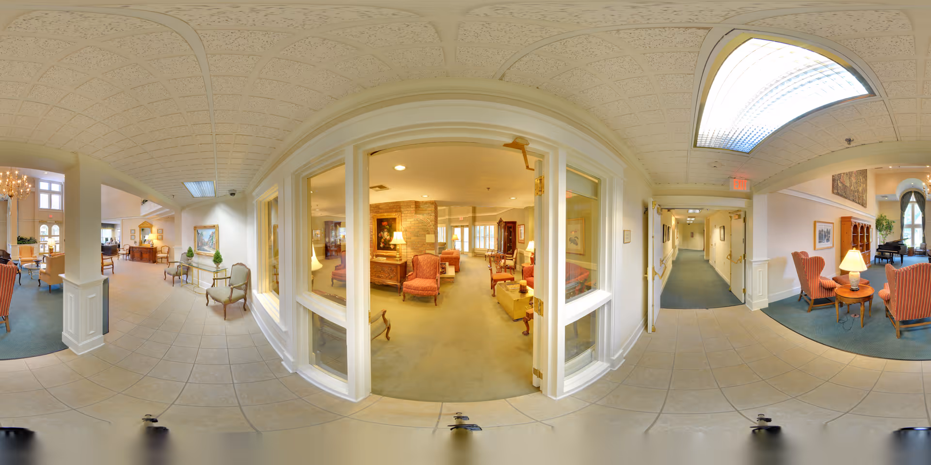 A spacious senior living facility interior with a tiled floor hallway leading to multiple seating areas furnished with armchairs, side tables, lamps, and decorative plants. The area includes a carpeted lounge with classic furniture and paintings on the walls. Large windows and glass doors provide a view into an adjacent room with more seating and warm lighting. The ceiling has a patterned design with recessed lighting and a skylight.