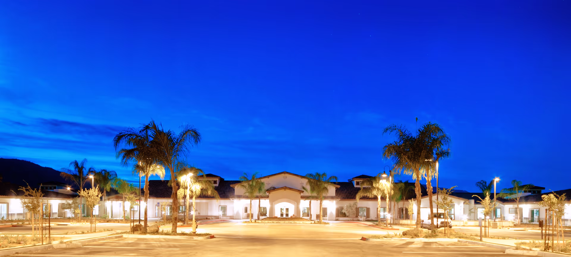 Wide exterior view of Manzanita Village senior living facility at dusk, showing a single-story building with a tiled roof, illuminated by outdoor lights and surrounded by palm trees and a spacious parking area.