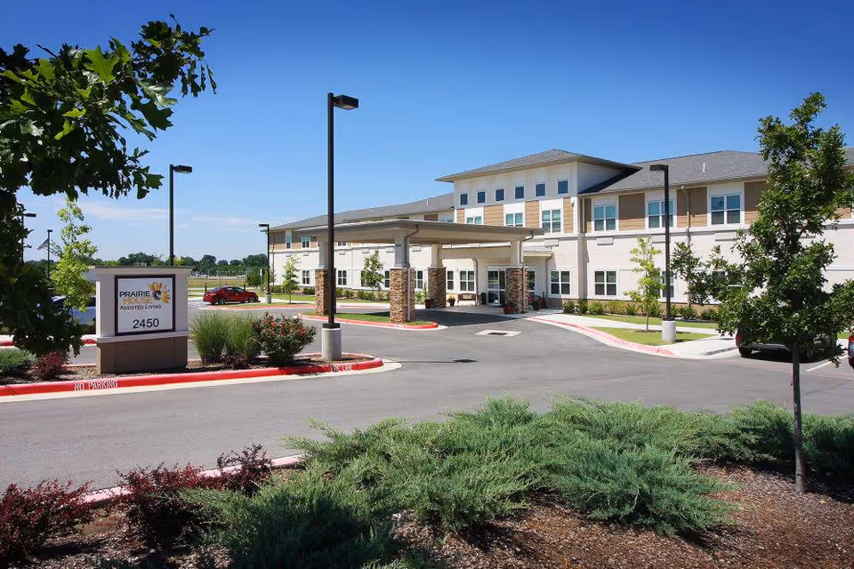 Exterior view of Prairie House Assisted Living facility showing a two-story building with a covered entrance, surrounded by landscaped greenery, trees, and a clear blue sky.