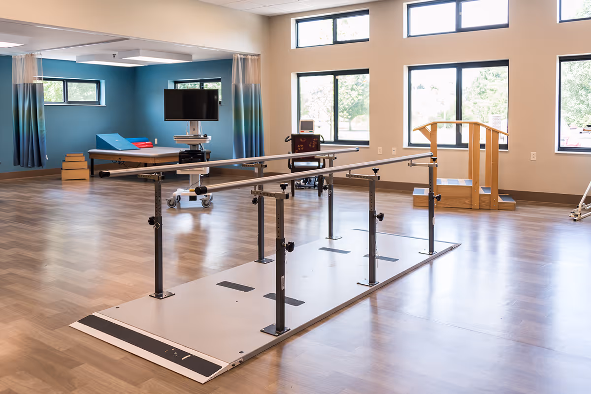 A spacious rehabilitation room with parallel bars for walking exercises, a therapy bed, a wheelchair, a TV on a mobile stand, and a wooden staircase for physical therapy. Large windows allow natural light to fill the room.