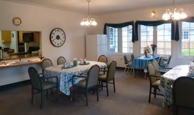 A dining area in an assisted living facility with three tables covered in patterned tablecloths, each surrounded by chairs. The room has large windows with dark valances, a white refrigerator, and a wall clock. A kitchen pass-through window is visible on the left side.