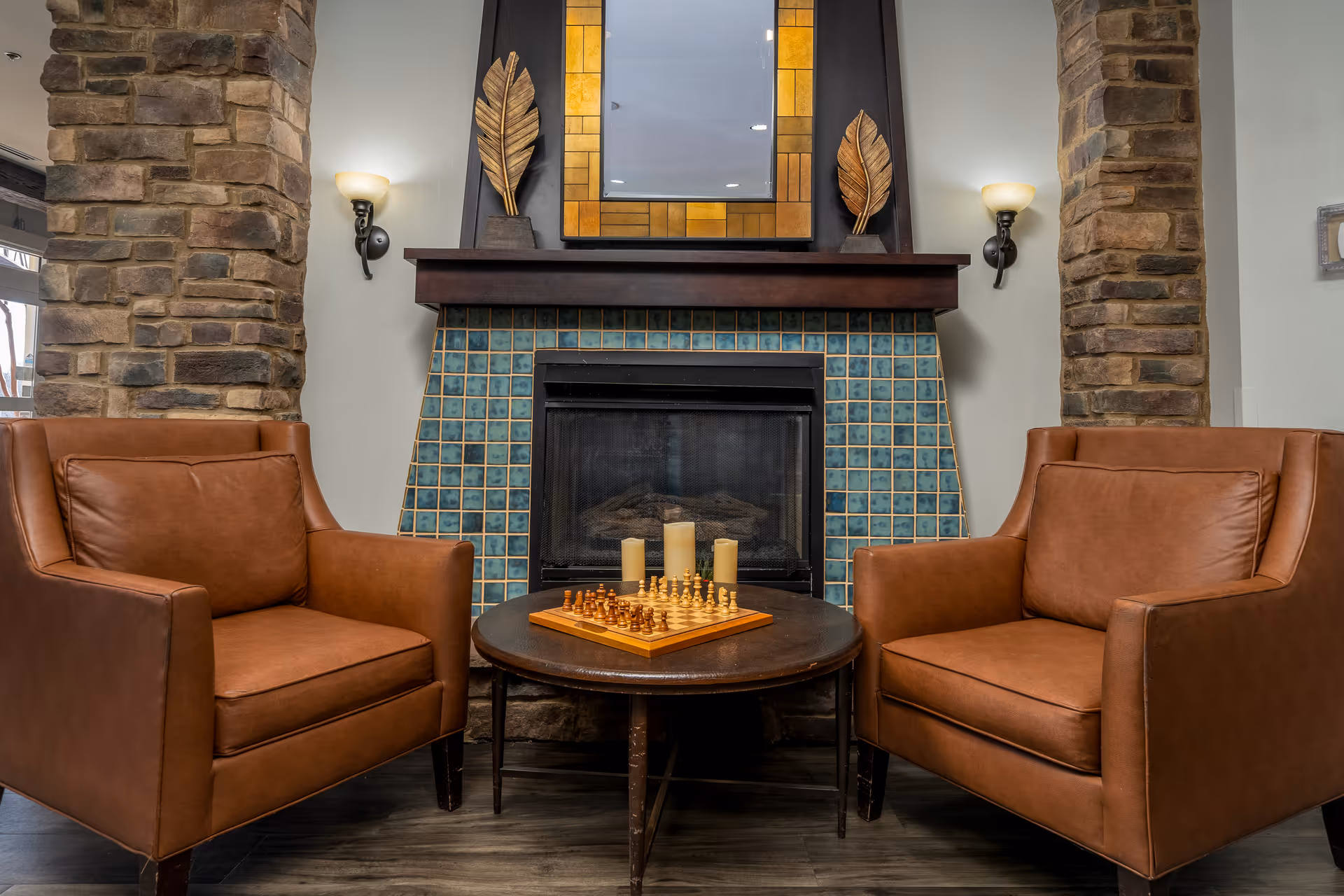 Two brown leather armchairs facing each other with a round wooden table between them holding a chessboard and three candles, set in front of a fireplace with blue-green tiles and stone pillars on either side. Above the fireplace is a mirror framed with yellow tiles and decorative leaf sculptures on each side, with wall sconces providing light.