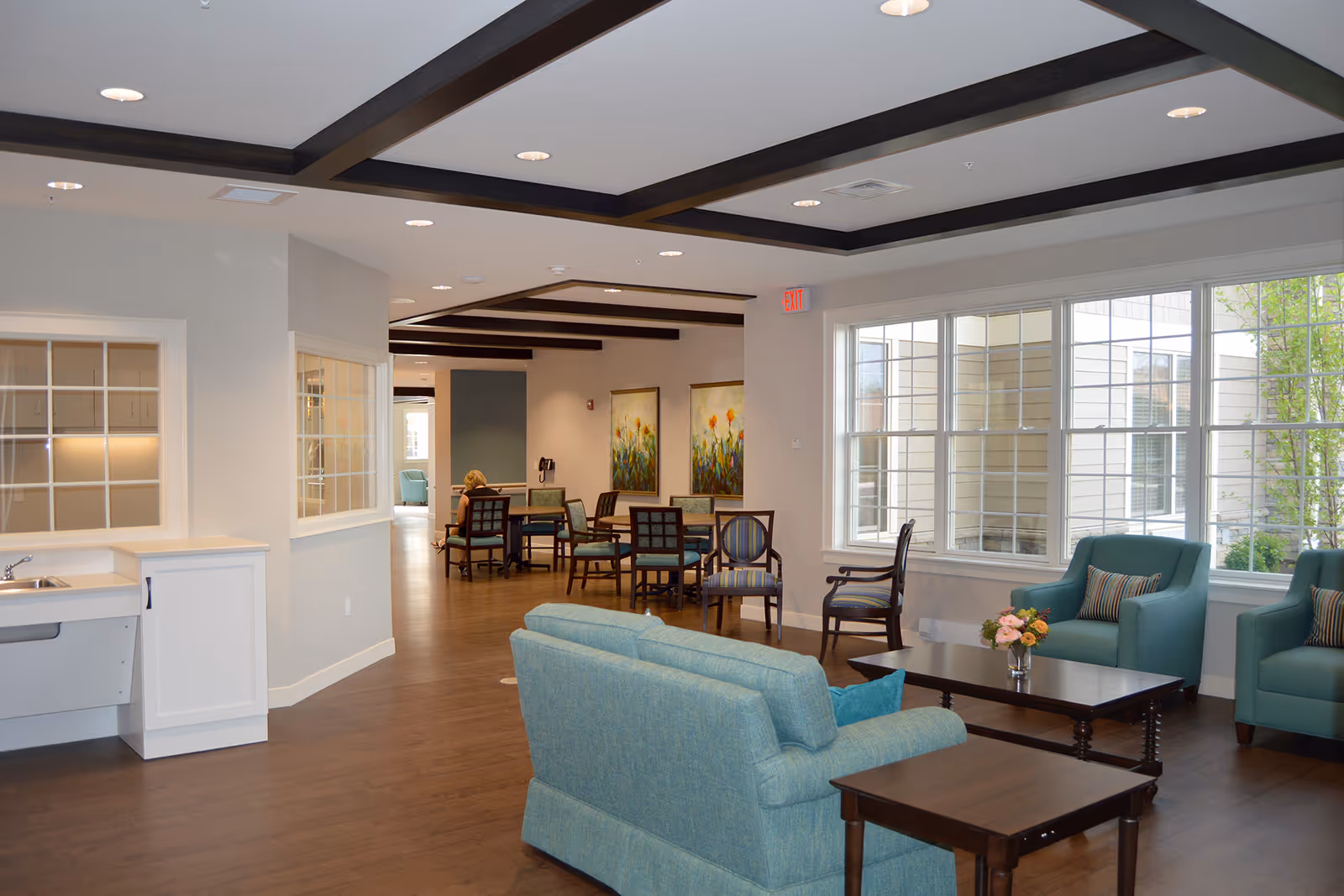 Bright communal living room/lounge with teal sofas and armchairs, coffee tables, dining tables in the background, and large windows.