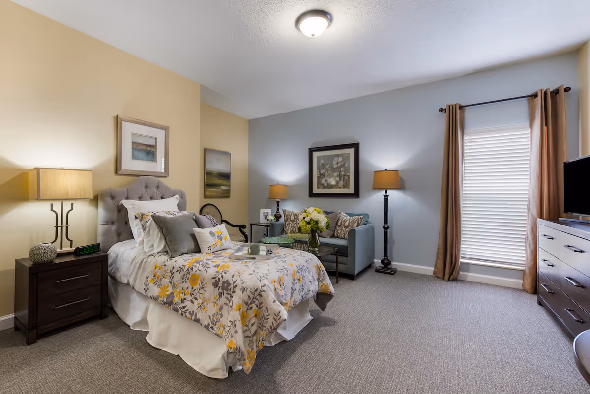 A cozy bedroom in Arbor Terrace Knoxville featuring a single bed with floral bedding, a nightstand with a lamp and alarm clock, a small sofa with decorative pillows, two floor lamps, framed artwork on the walls, a window with brown curtains and blinds, and a dresser with a TV on top.