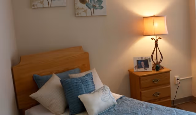 A cozy bedroom with a wooden headboard bed made with blue and white pillows and a blue quilt. Next to the bed is a wooden nightstand with two drawers, a decorative lamp, and a framed photo. Two floral paintings hang on the beige wall above the bed.